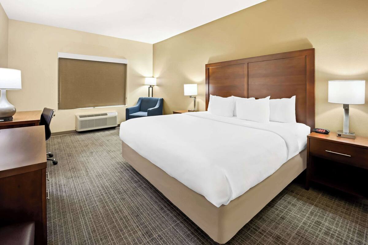 Une chambre d’hôtel avec un grand lit, un bureau avec une chaise, une lampe de bureau, une table de chevet avec une lampe et un climatiseur fixé au mur.