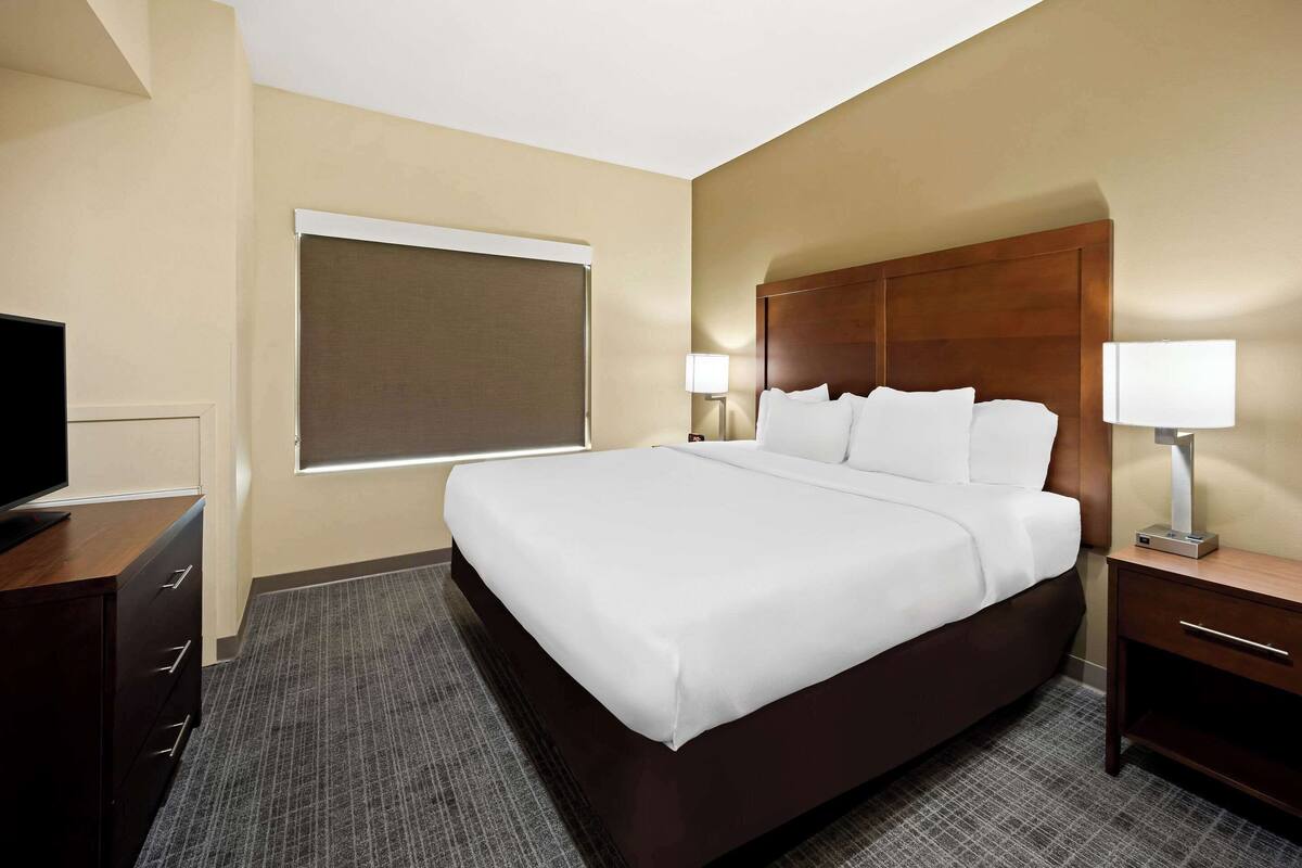 Une chambre d’hôtel avec un grand lit, deux tables de chevet avec des lampes, un téléviseur posé sur une commode et une fenêtre avec des stores.