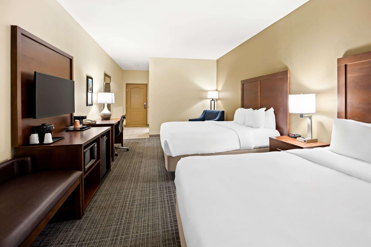 A hotel room with two beds, a TV, a desk with a lamp, and a door in the background.