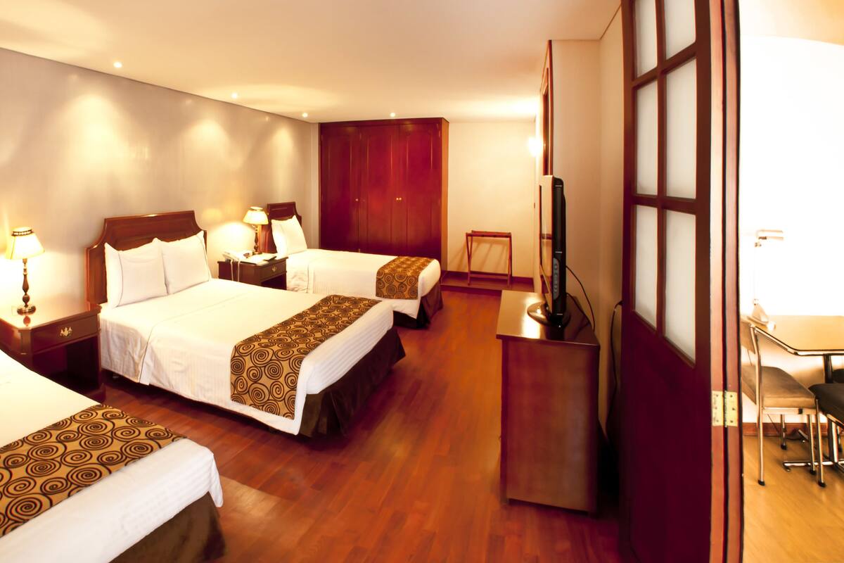 A hotel room with three beds, wooden flooring, a TV, and a desk with chairs.