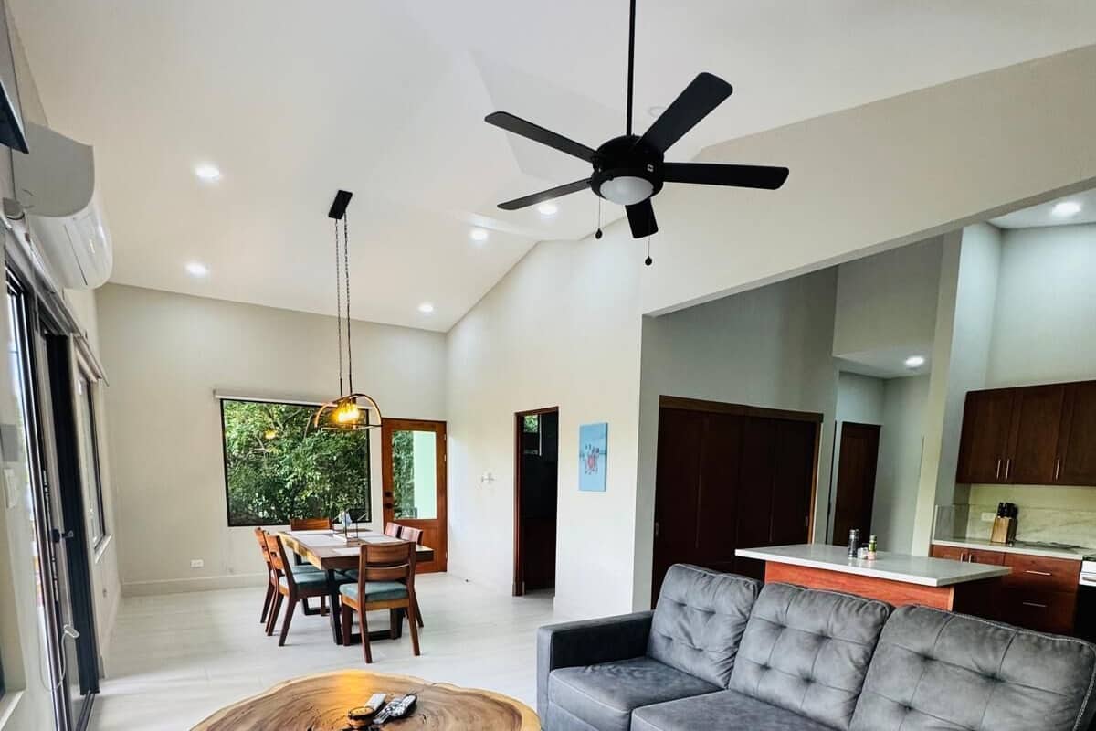 A modern living room with a sectional sofa, a unique wooden coffee table, a ceiling fan, and a dining area with a view of greenery.