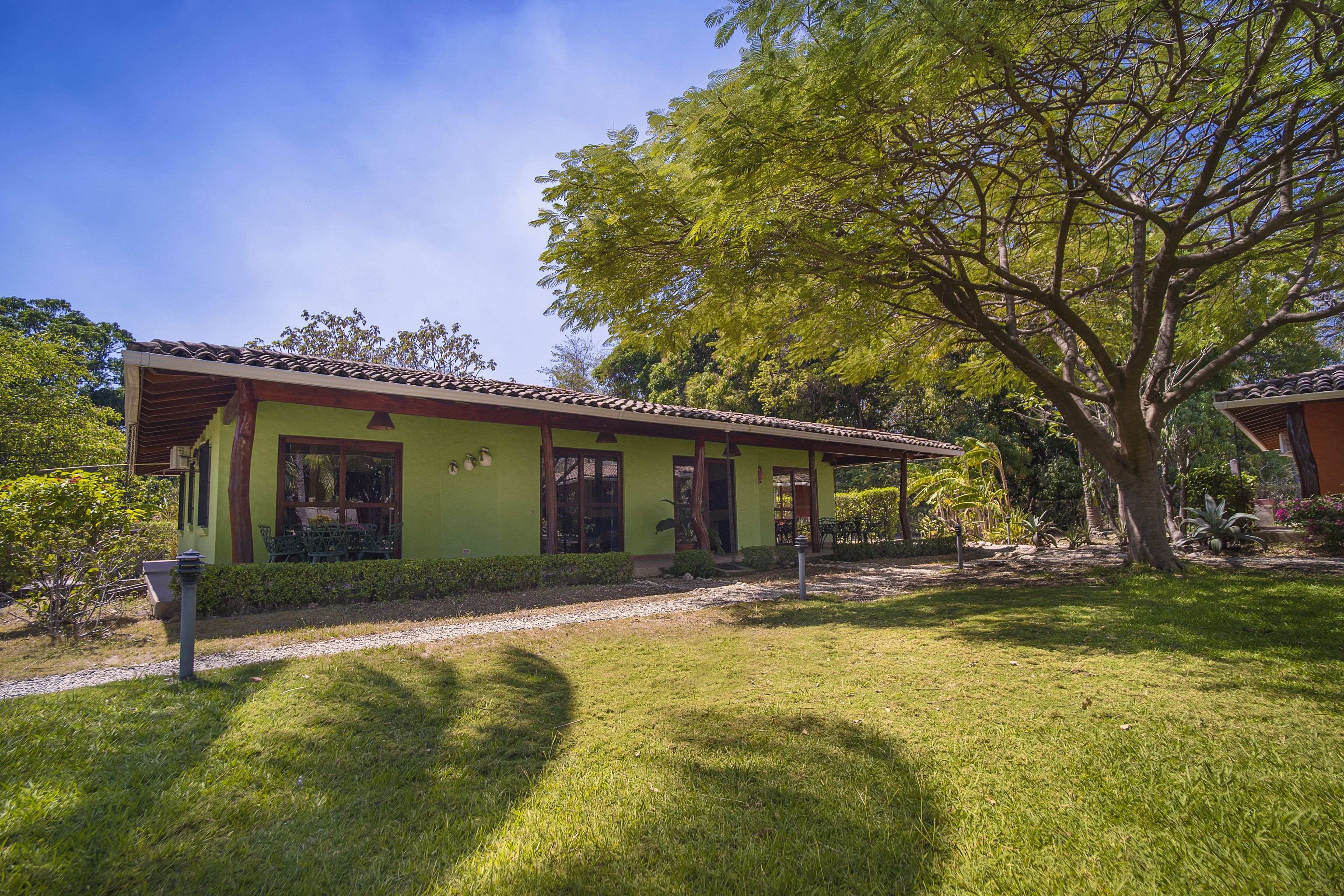 A green building with a tiled roof, surrounded by a well-maintained lawn and a large tree.