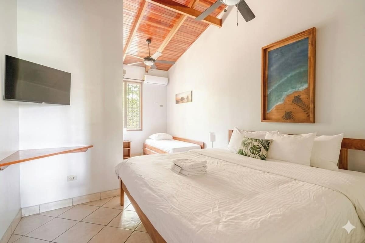 A bedroom with a bed, a TV mounted on the wall, a ceiling fan, and a painting of a beach scene.