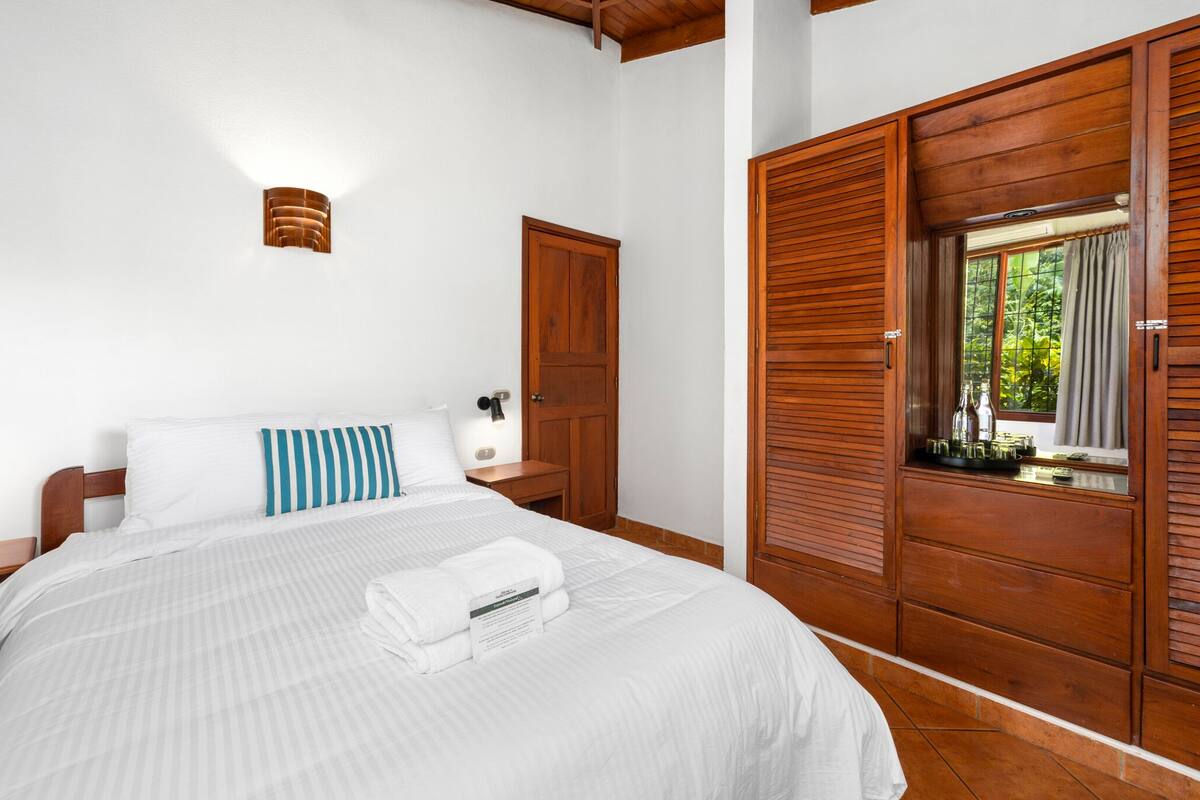 A neatly made bed with white linens and a blue and white striped pillow, a bedside table with a lamp, and a wooden wardrobe with a window view.