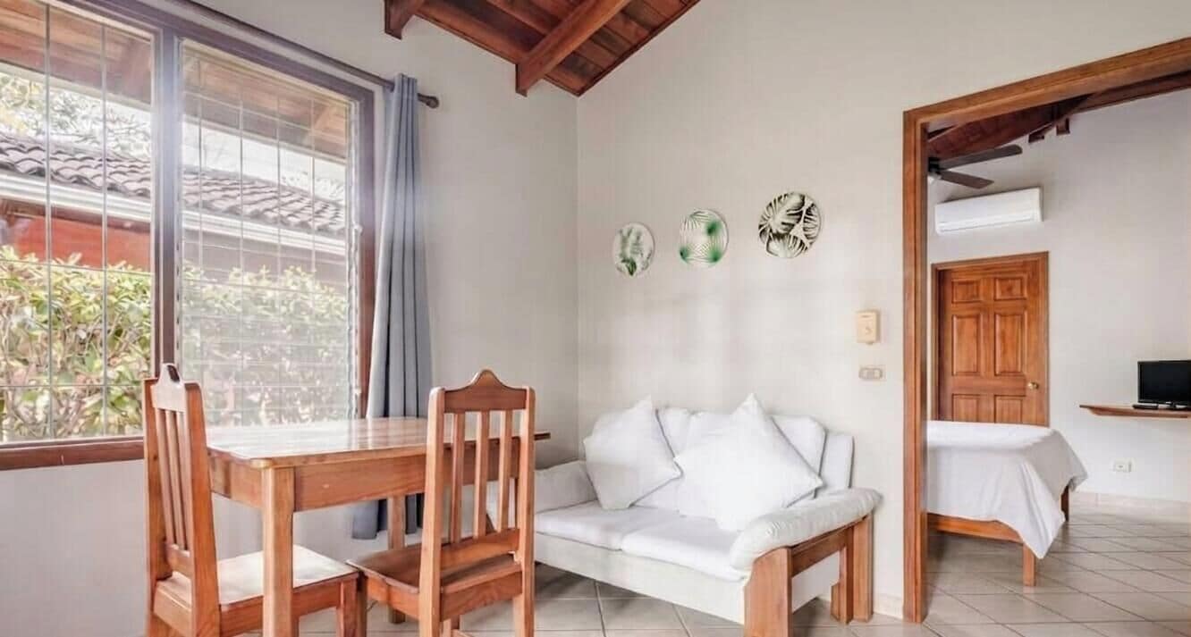 A room with a wooden dining table, a white sofa, a small bed, and a window with a view of greenery.