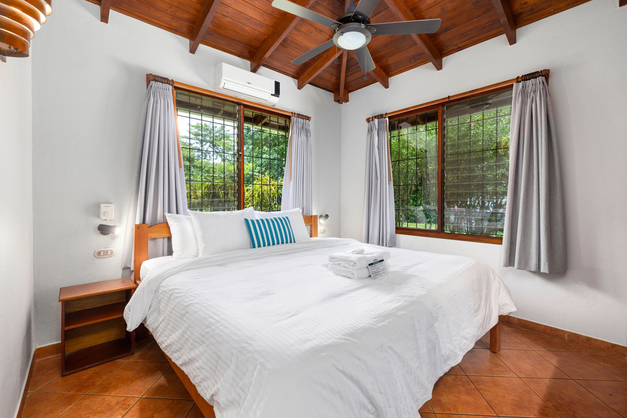 A bedroom with a large bed, wooden headboard, and a ceiling fan. There are two windows with curtains, and an air conditioning unit on the wall.