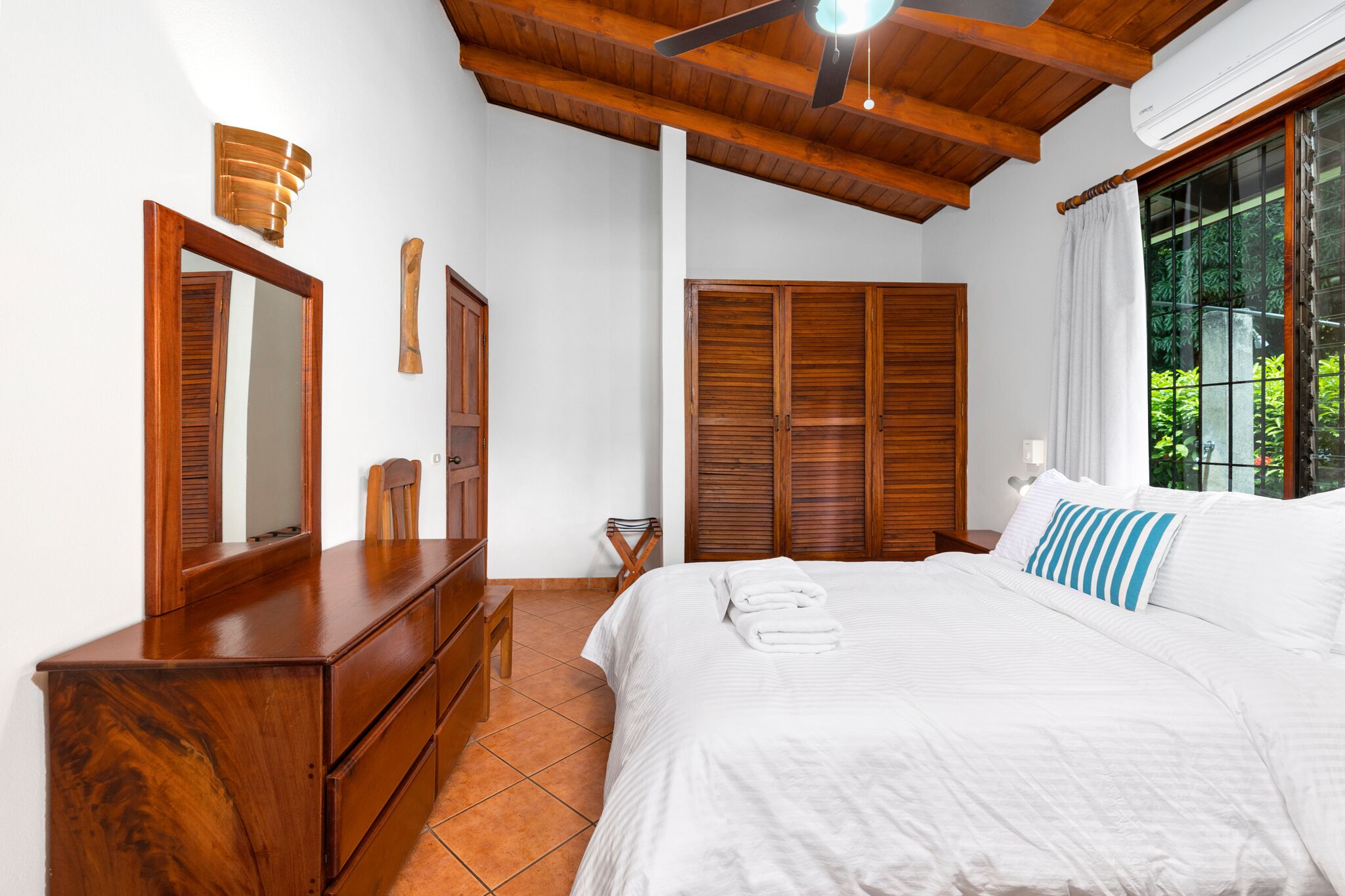 Een slaapkamer met een houten dressoir, een bed met wit beddengoed, een plafondventilator en een raam met uitzicht op groen.