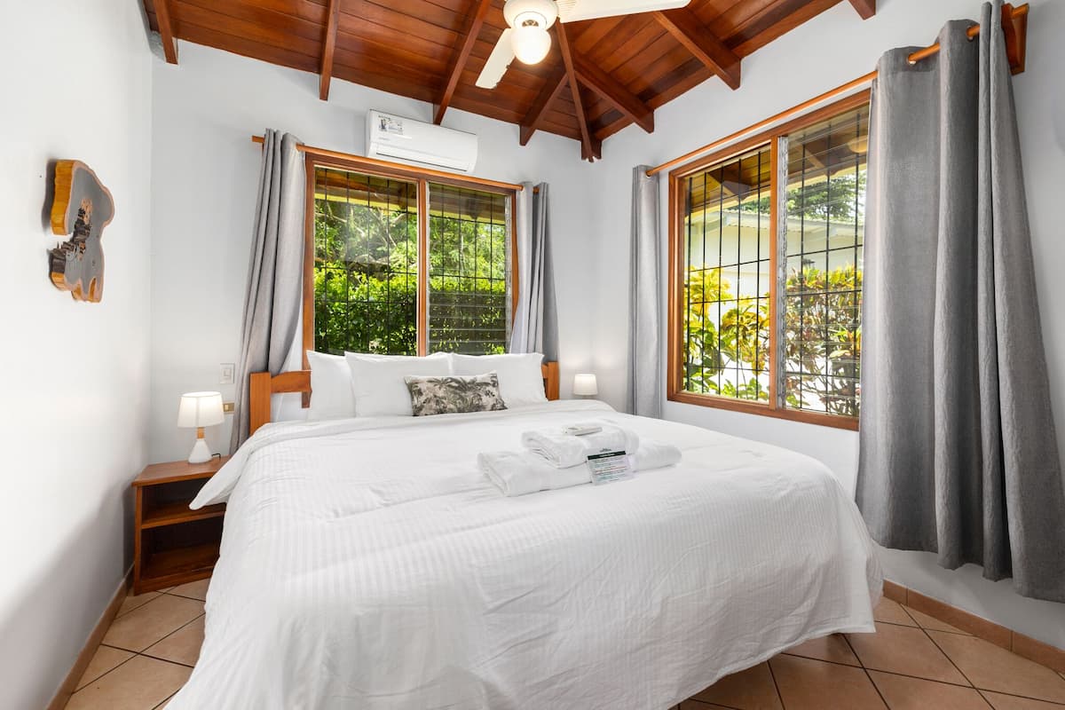 A hotel room with a large bed, wooden headboard, two bedside tables with lamps, a ceiling fan, and a window with curtains.