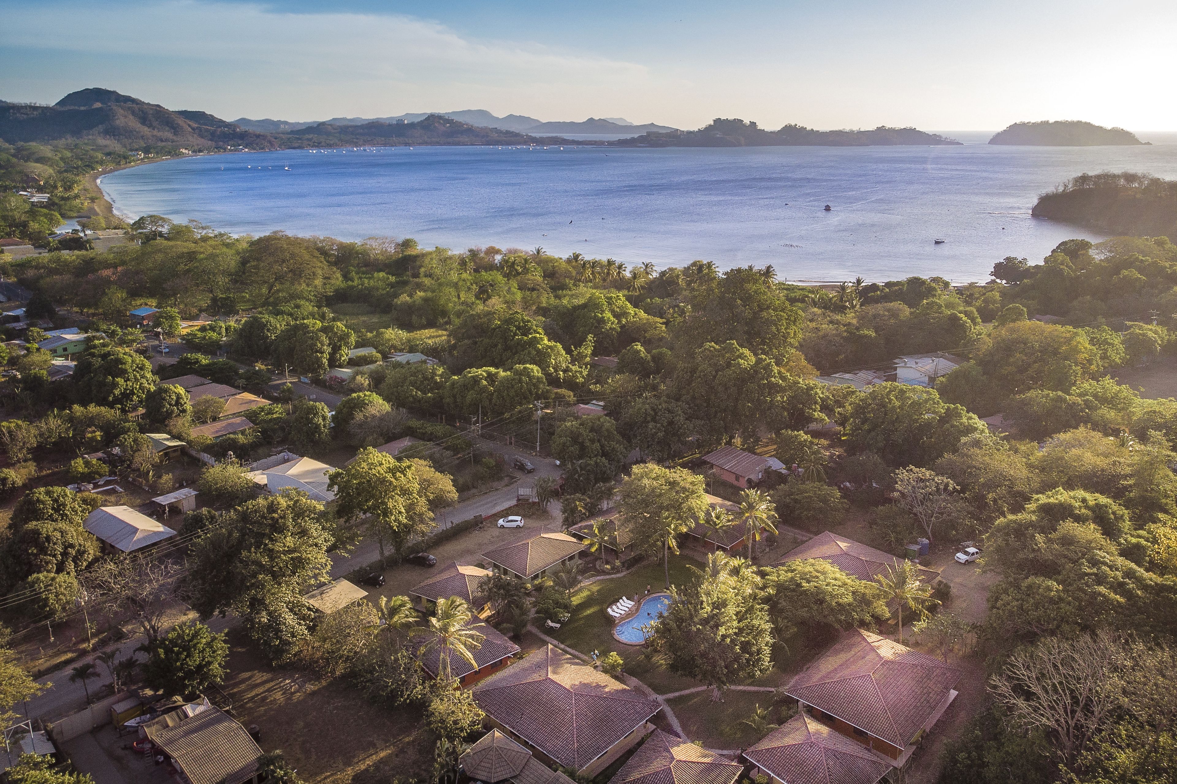 An aerial view of a coastal residential area with houses, a swimming pool, and a large body of water.