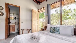 A bedroom with a bed, a bedside table, a window with a view of greenery, and a bathroom visible through an open door. - Bahia Esmeralda Boutique Hotel & Villas (Tempate)