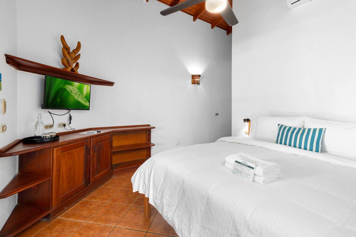 A hotel room with a wooden desk, a flat-screen TV, a bed with white bedding, and a ceiling fan.