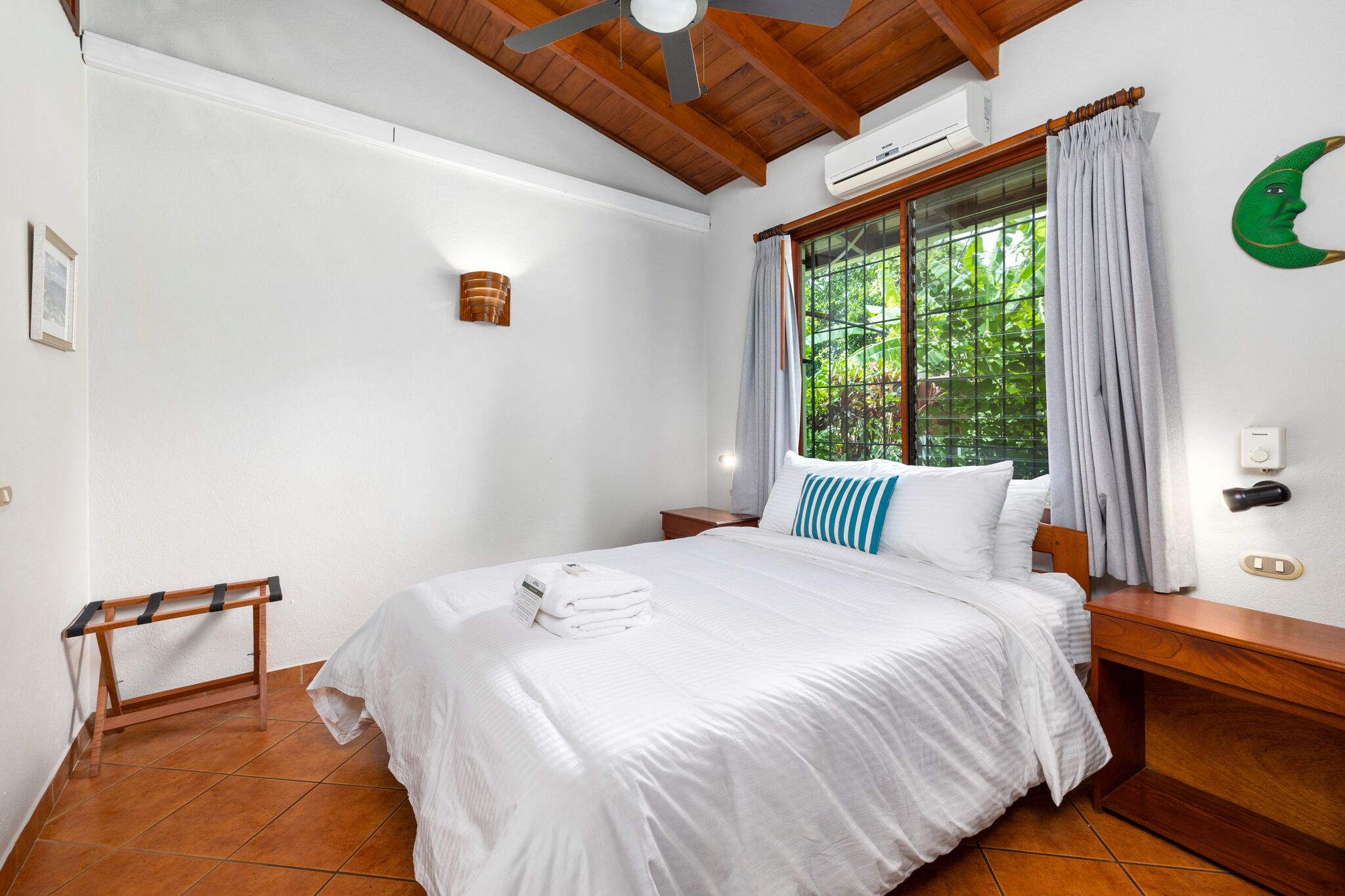 A clean, well-lit hotel room with a large bed, a wooden headboard, a window with curtains, and a ceiling fan.