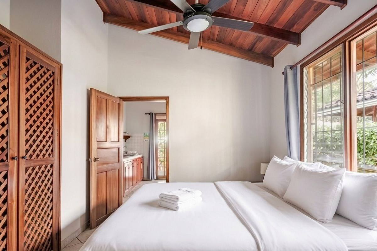 A bedroom with a bed, wooden wardrobe, ceiling fan, and large windows.