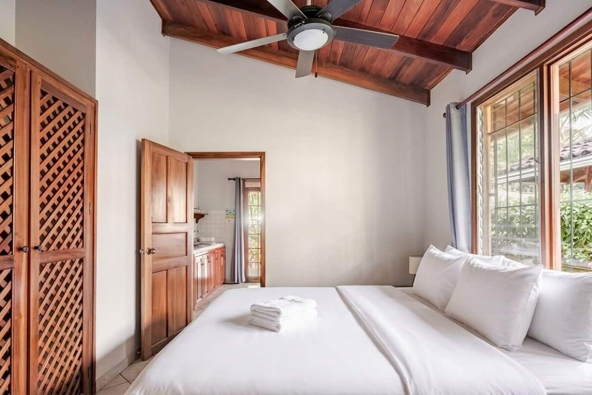 A bedroom with a bed, wooden wardrobe, ceiling fan, and large windows.