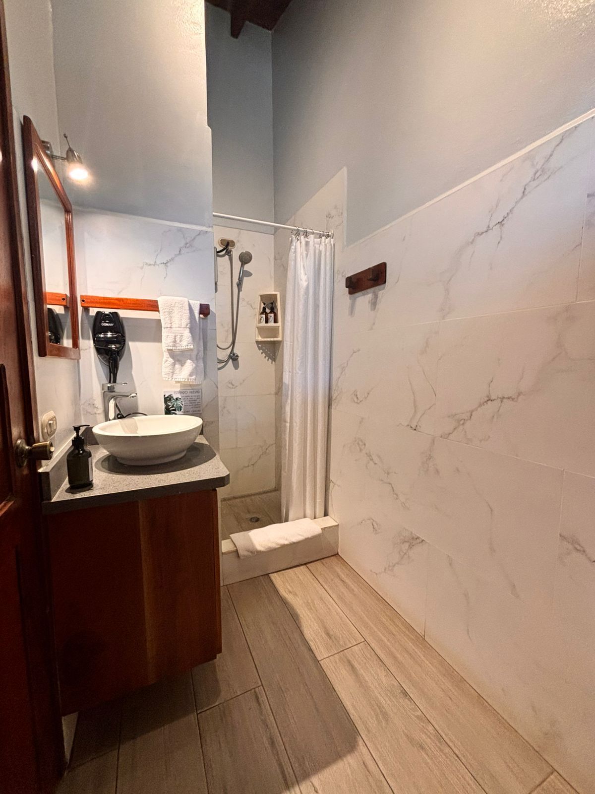 Un baño con pared de mármol, tocador de madera, lavamanos, ducha y espejo.