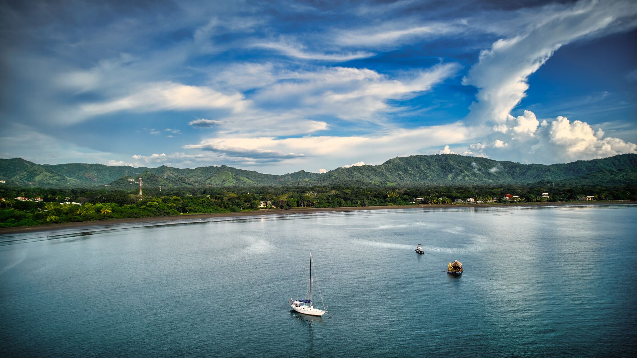 A calm bay with a sailboat and a motorboat, surrounded by lush green hills and a clear sky.