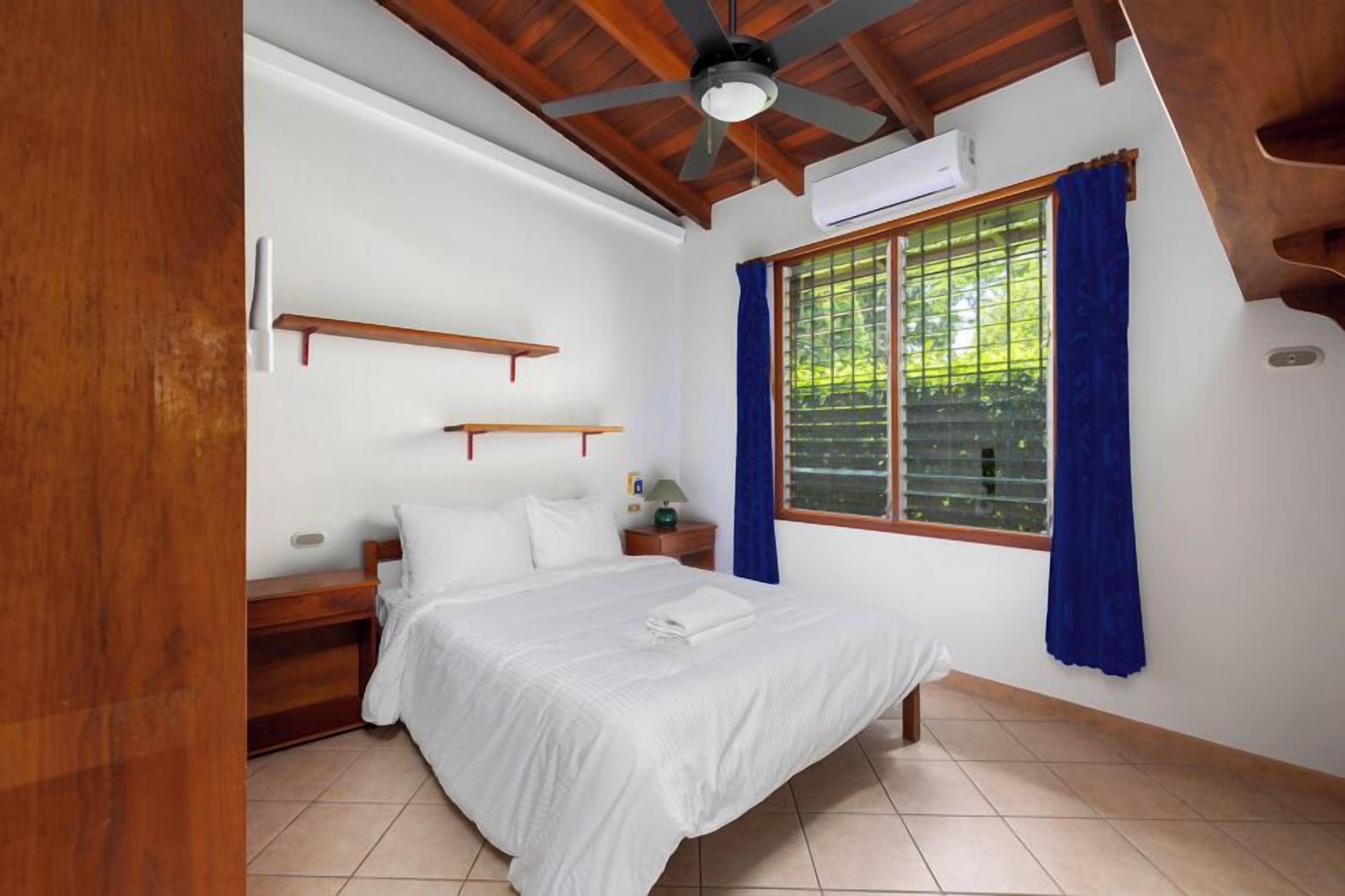 A bedroom with a bed, a nightstand, a window with blue curtains, and a ceiling fan.