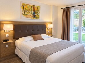 Une chambre d’hôtel avec un grand lit, deux lampes de chevet, un tableau représentant une scène de forêt et une vue sur l’extérieur par une fenêtre avec des rideaux.