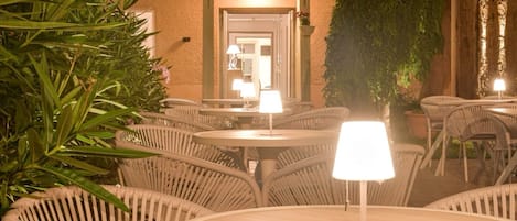 An outdoor patio with wicker chairs and tables, a building in the background, and a potted plant.