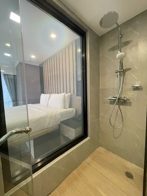 A modern bathroom with a glass shower enclosure, a walk-in shower, and a visible bedroom with a bed and curtains. - Citin Pratunam Bangkok Hotel by Compass Hospitality (Bangkok)