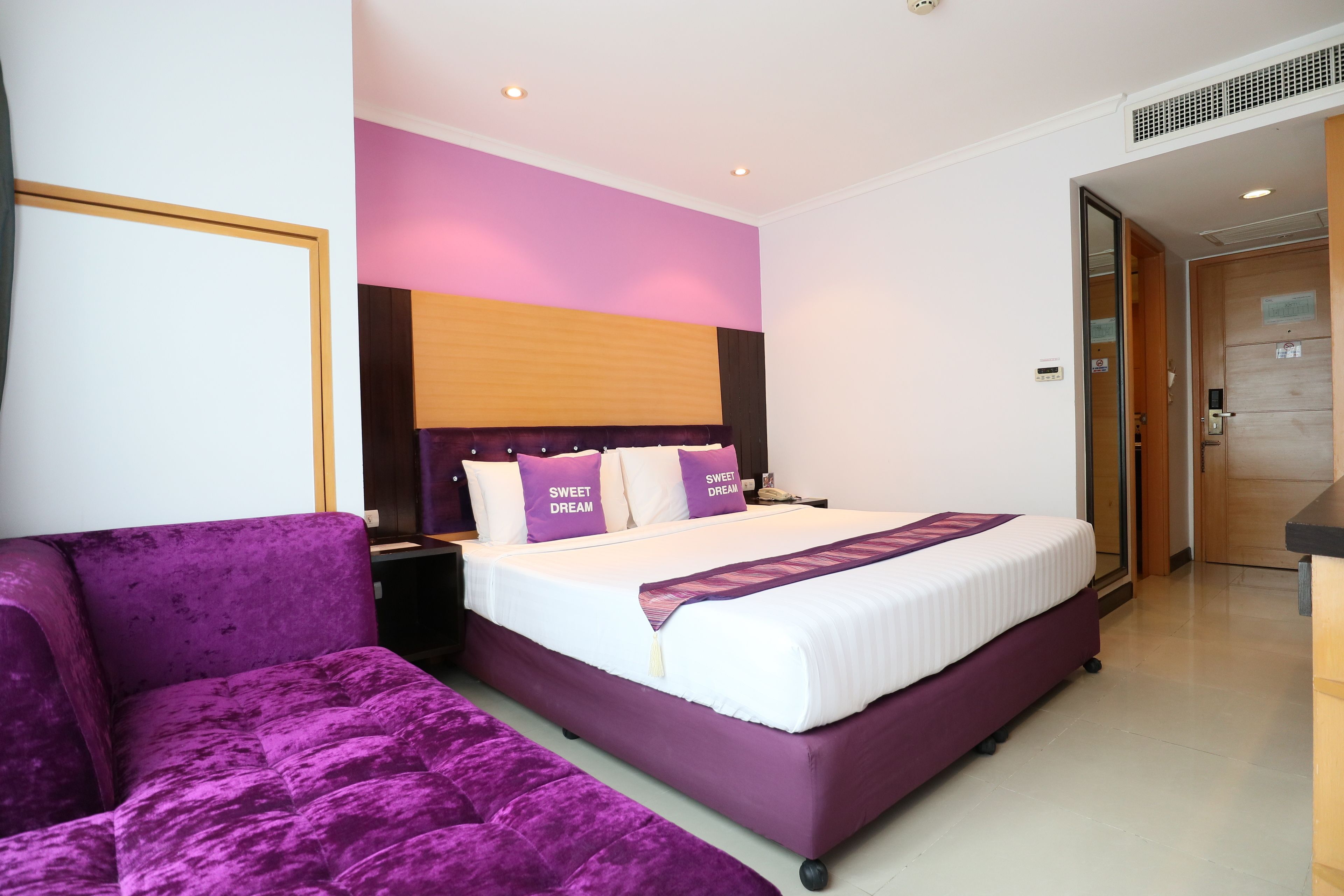 Une chambre d’hôtel moderne avec un grand lit, un canapé violet et une tête de lit en bois.