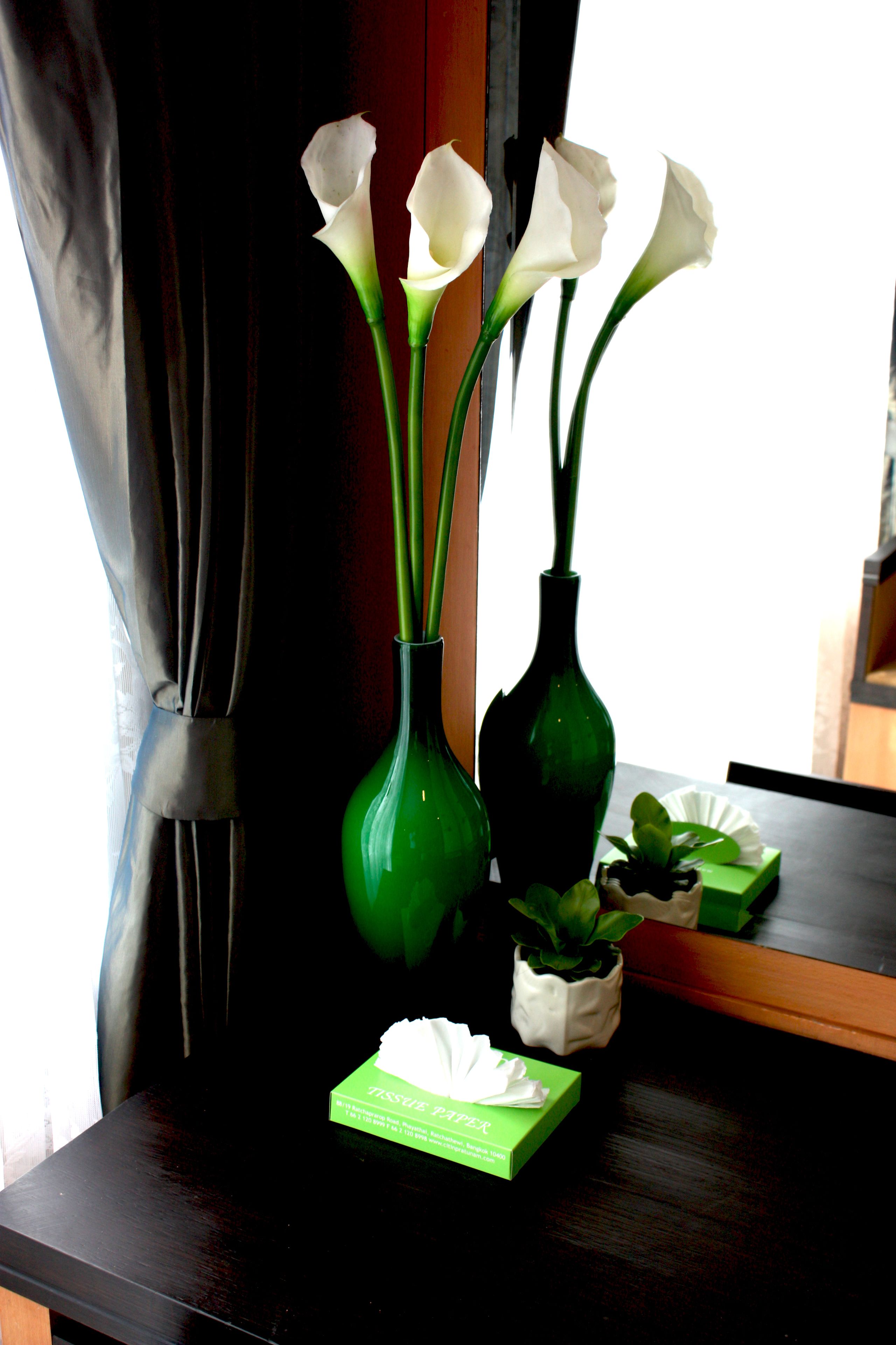 A window with white calla lilies in green vases, a tissue box, and a small potted plant on a dark wooden surface.