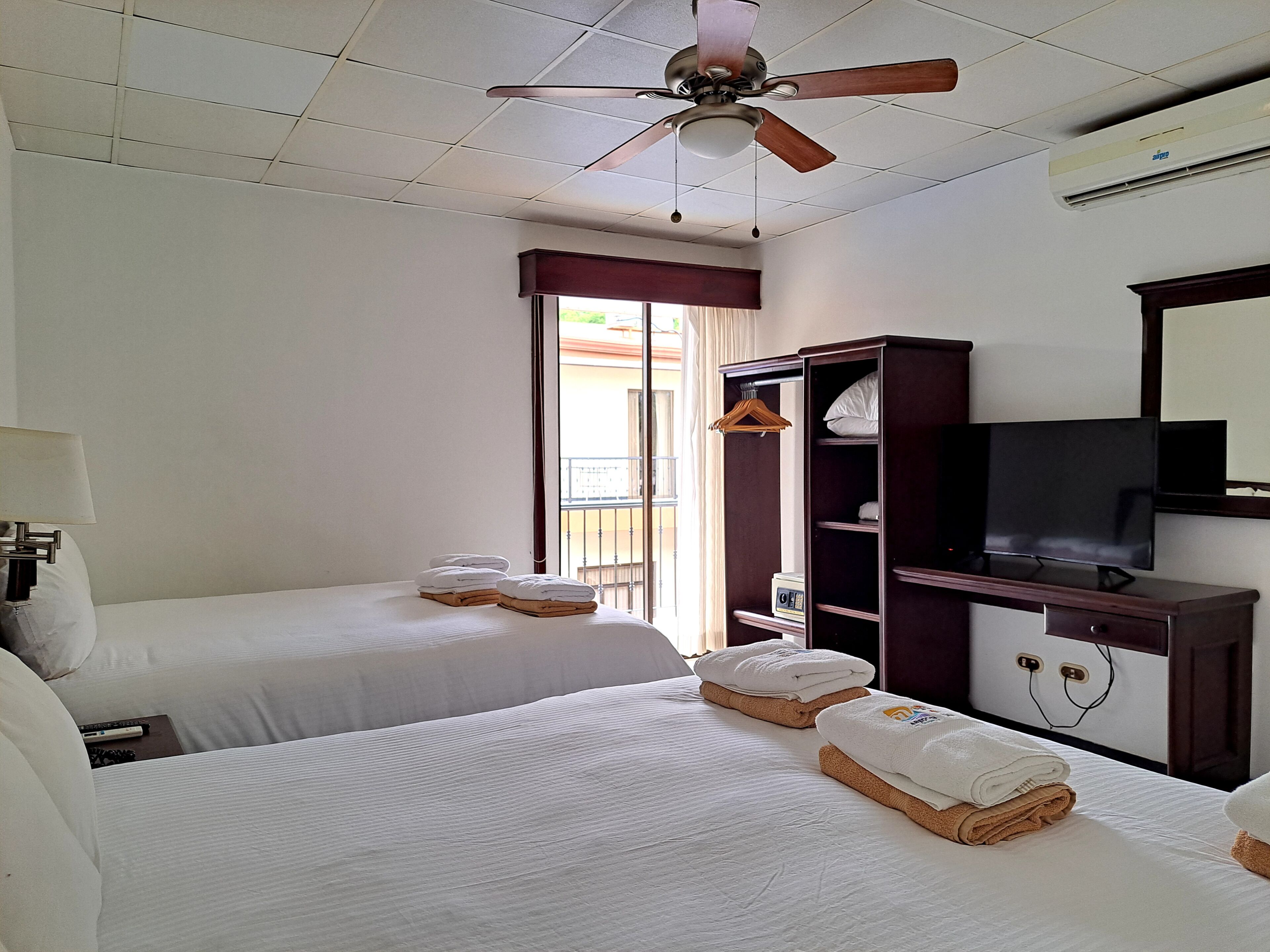 Habitación de hotel con dos camas, ventilador de techo, televisión, armario y ventana con vistas.