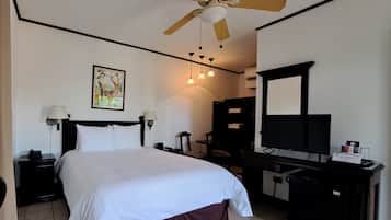 A hotel room with a bed, a desk, a chair, a ceiling fan, a wall-mounted TV, and a painting.