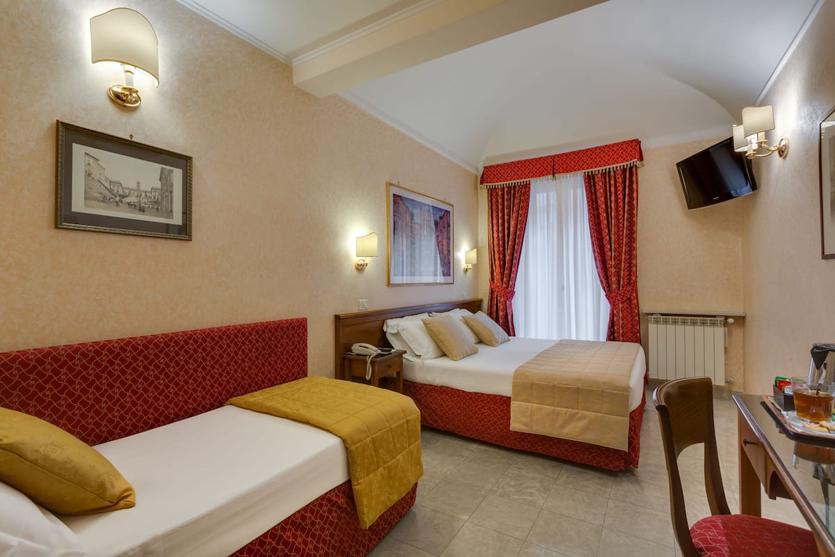 A hotel room with two beds, a desk, a chair, a TV, and a window with red curtains.