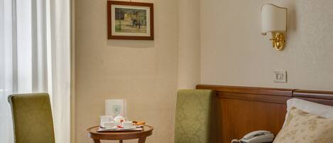 A hotel room with two green upholstered chairs, a small wooden table with a teapot and cups, a framed picture on the wall, a bedside lamp, and a telephone on the nightstand.