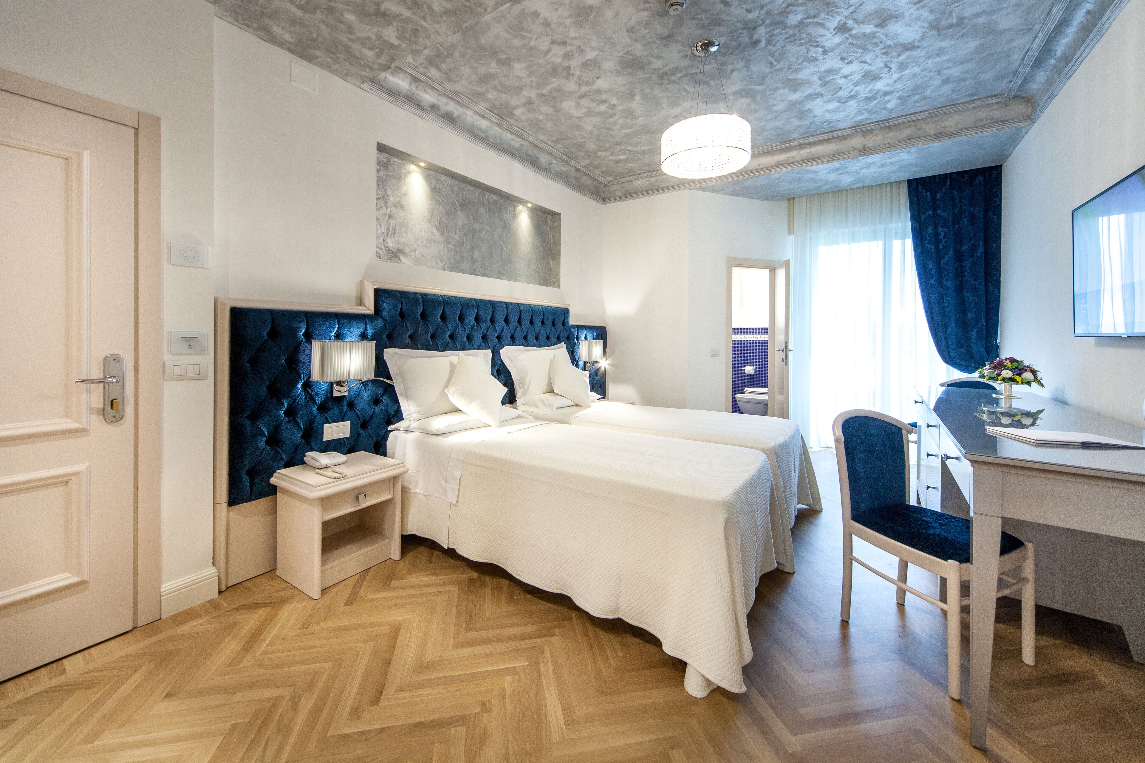 Una habitación de hotel con una cama grande, un escritorio y una silla. Hay una ventana con cortinas, una lámpara en el techo y una puerta.