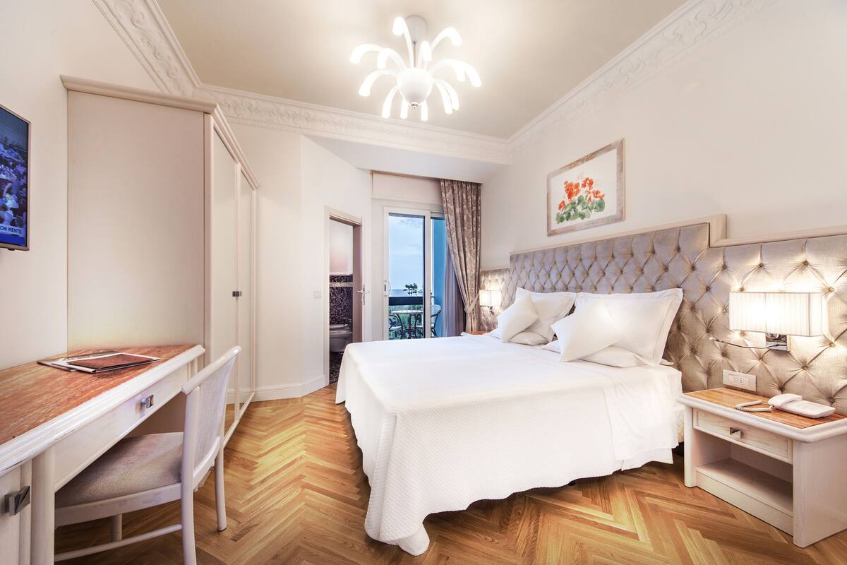 A hotel room with a large bed, a desk, and a chair. There is a window with curtains, a door leading to another room, and a wall-mounted telephone.
