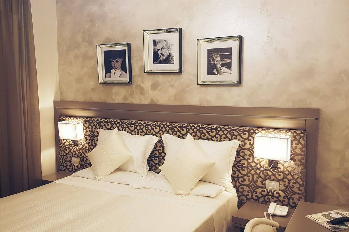A hotel room with a bed, two bedside lamps, a telephone, and three framed pictures on the wall.