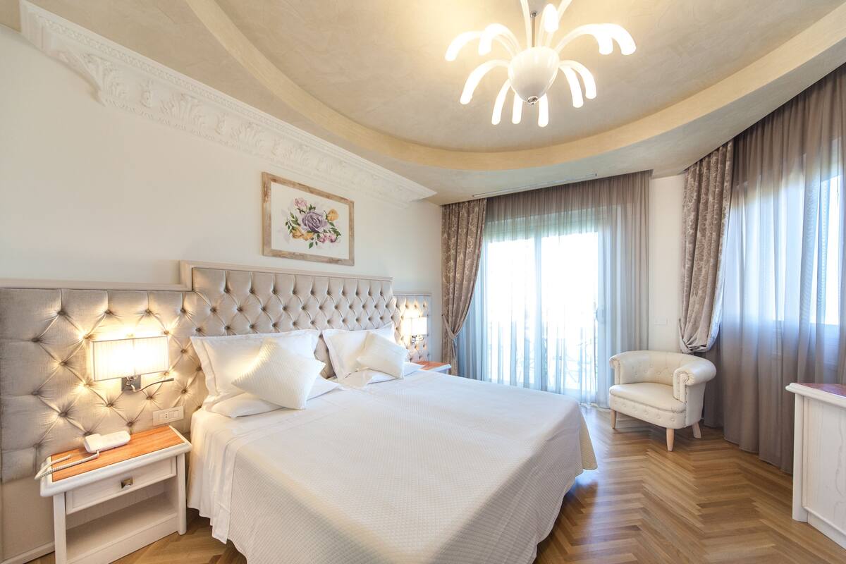 A hotel room with a large bed, a nightstand, a chair, a desk, and a chandelier.