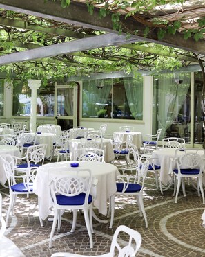 Une terrasse extérieure avec des tables et des chaises blanches, abritée par une pergola couverte de plantes grimpantes.