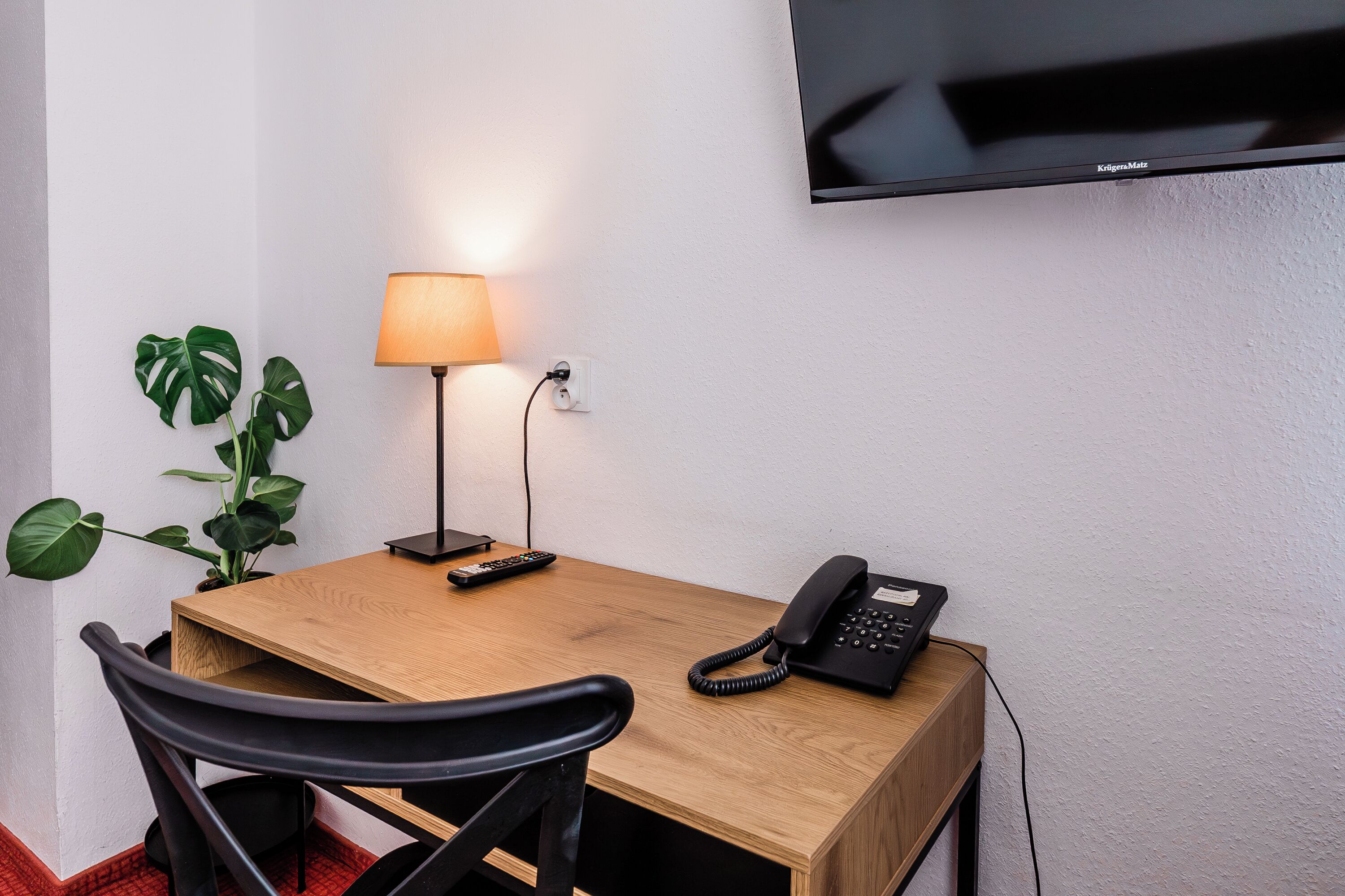 Una camera d'albergo con una scrivania in legno, una lampada, un telefono, un telecomando e una televisione a parete.