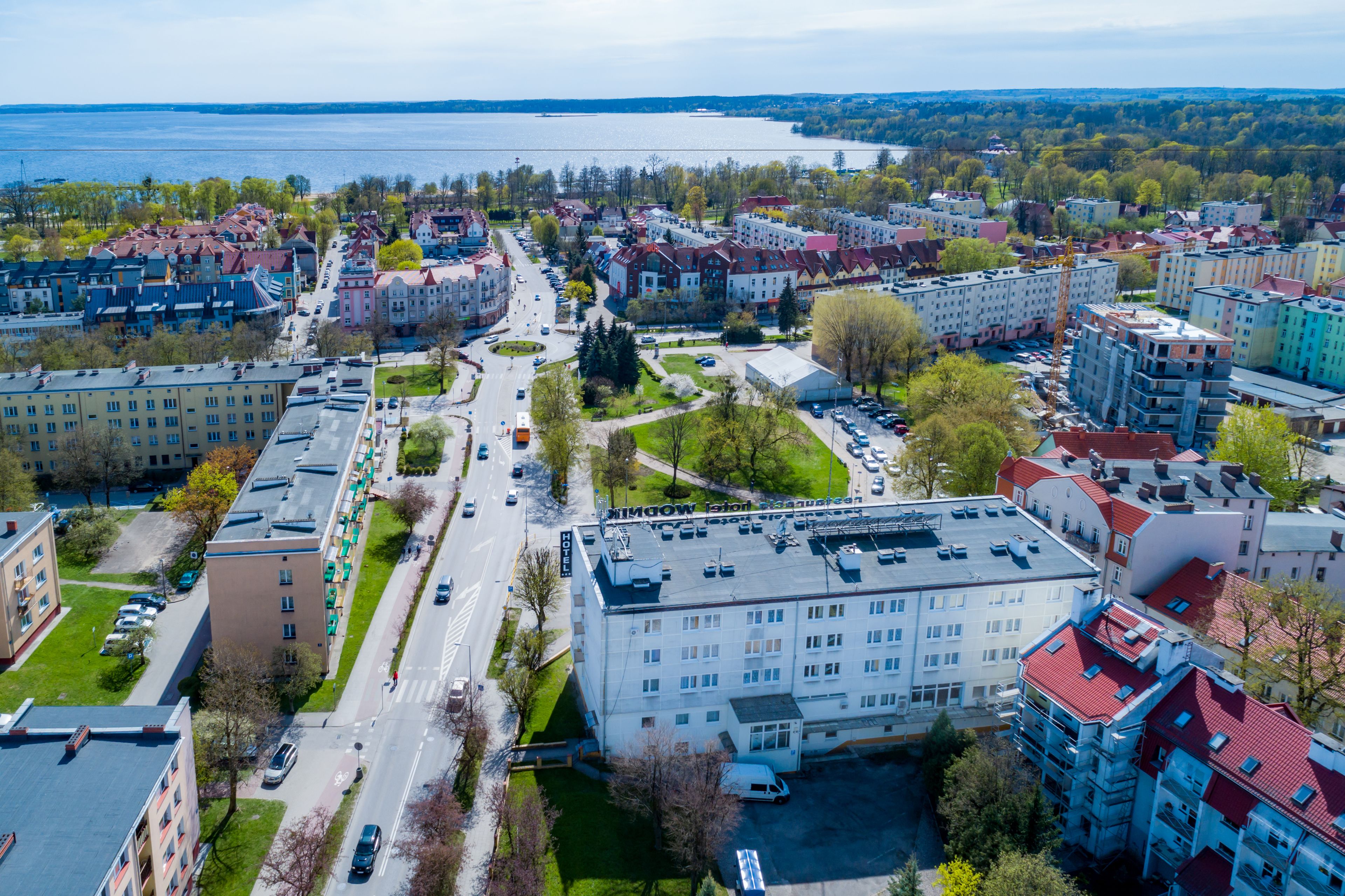 A multi-story building with a flat roof, surrounded by a mix of residential and commercial buildings, a main road, and a body of water in the background.