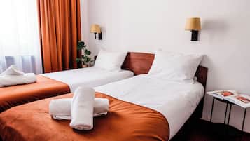 Ein Hotelzimmer mit zwei Betten, jeweils mit brauner Kopfstütze, weißer Bettwäsche und einem aufgerollten weißen Handtuch. Zwei Wandlampen, ein Topf mit einer Pflanze und eine Zeitschrift auf einem kleinen Tisch sind ebenfalls vorhanden.