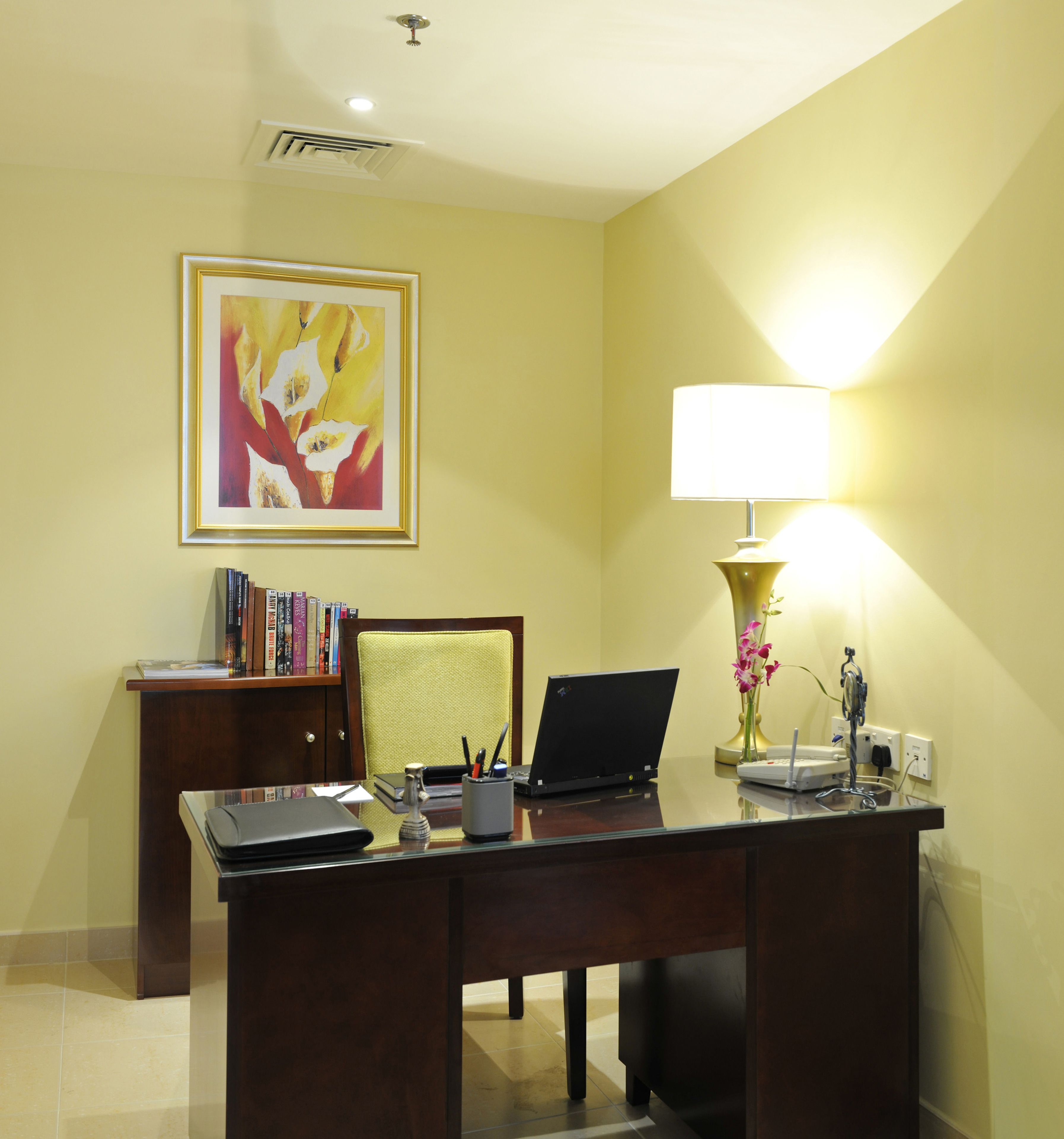 Een hotelkamer met een bureau, een laptop, een lamp, een boekenplank en een lijstje in een frame aan de muur.