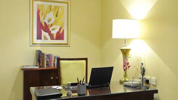 Een hotelkamer met een bureau, een laptop, een lamp, een boekenplank en een lijstje in een frame aan de muur.