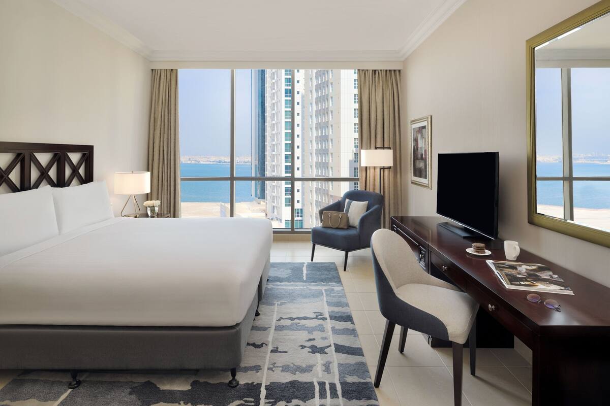 A hotel room with a large bed, a desk, a chair, a TV, and a view of the city and sea.
