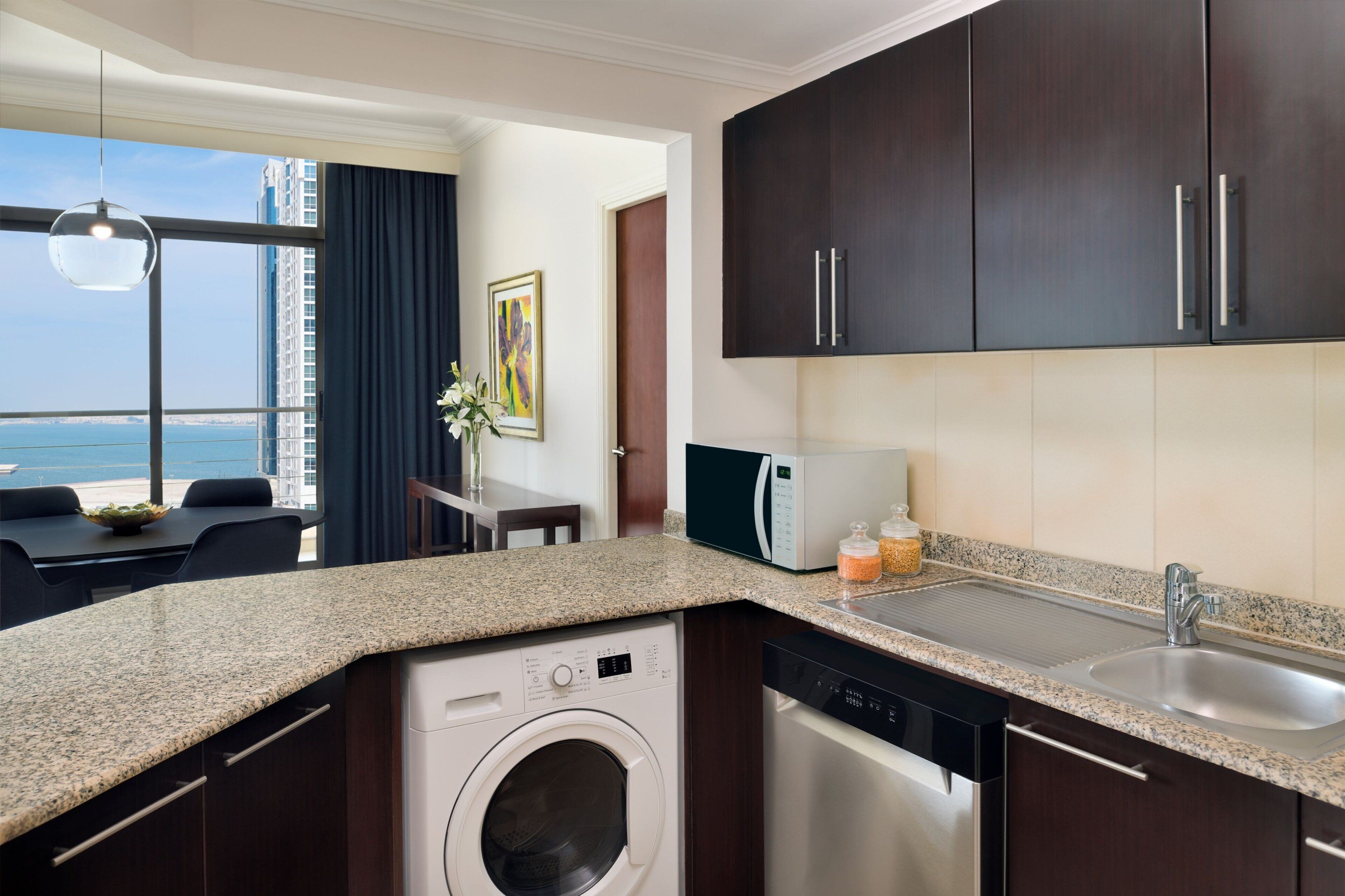 A modern kitchen with granite countertops, a built-in washer and dishwasher, dark wooden cabinets, a stainless steel sink, and a view of the ocean through large windows.
