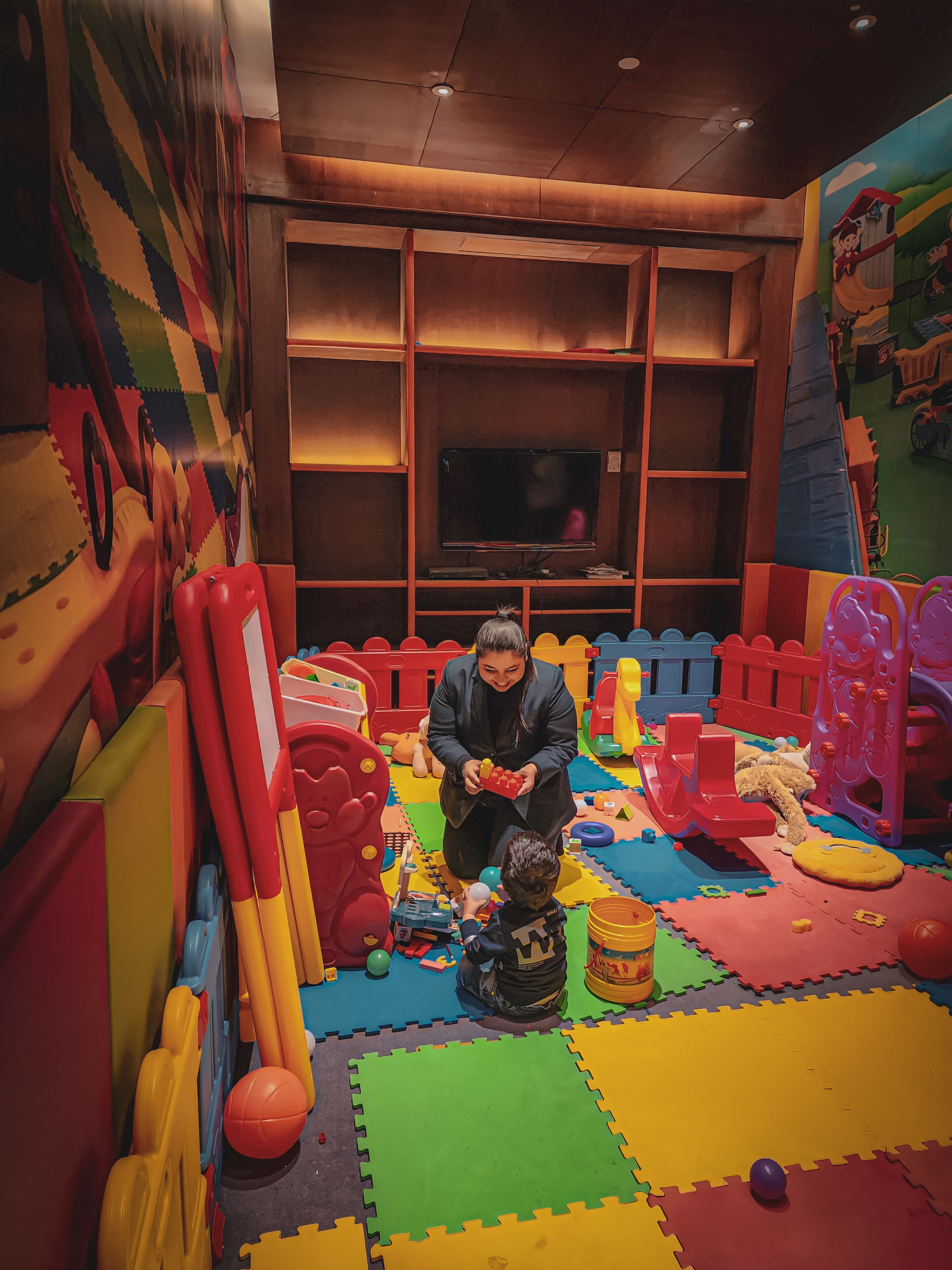 Zona de juegos con coloridas alfombras de espuma, juguetes y un televisor al fondo.