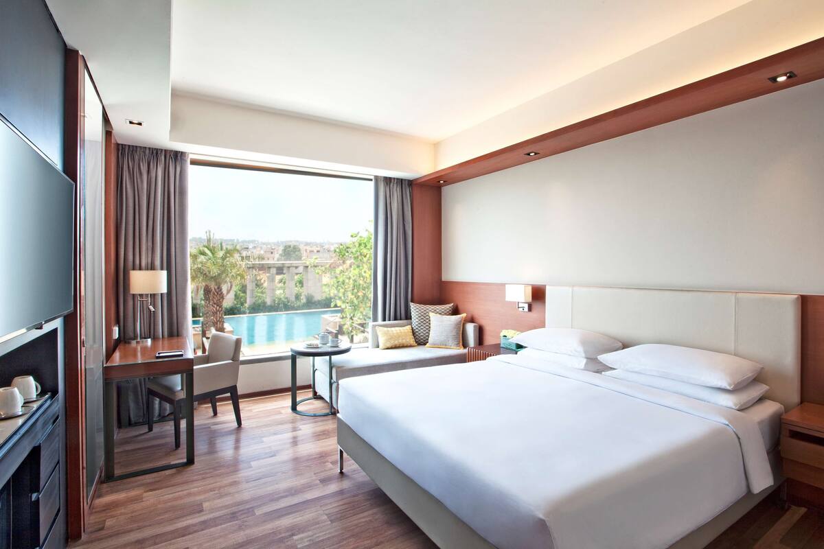A hotel room with a large bed, a desk with a chair, a TV, and a view of a swimming pool and cityscape.