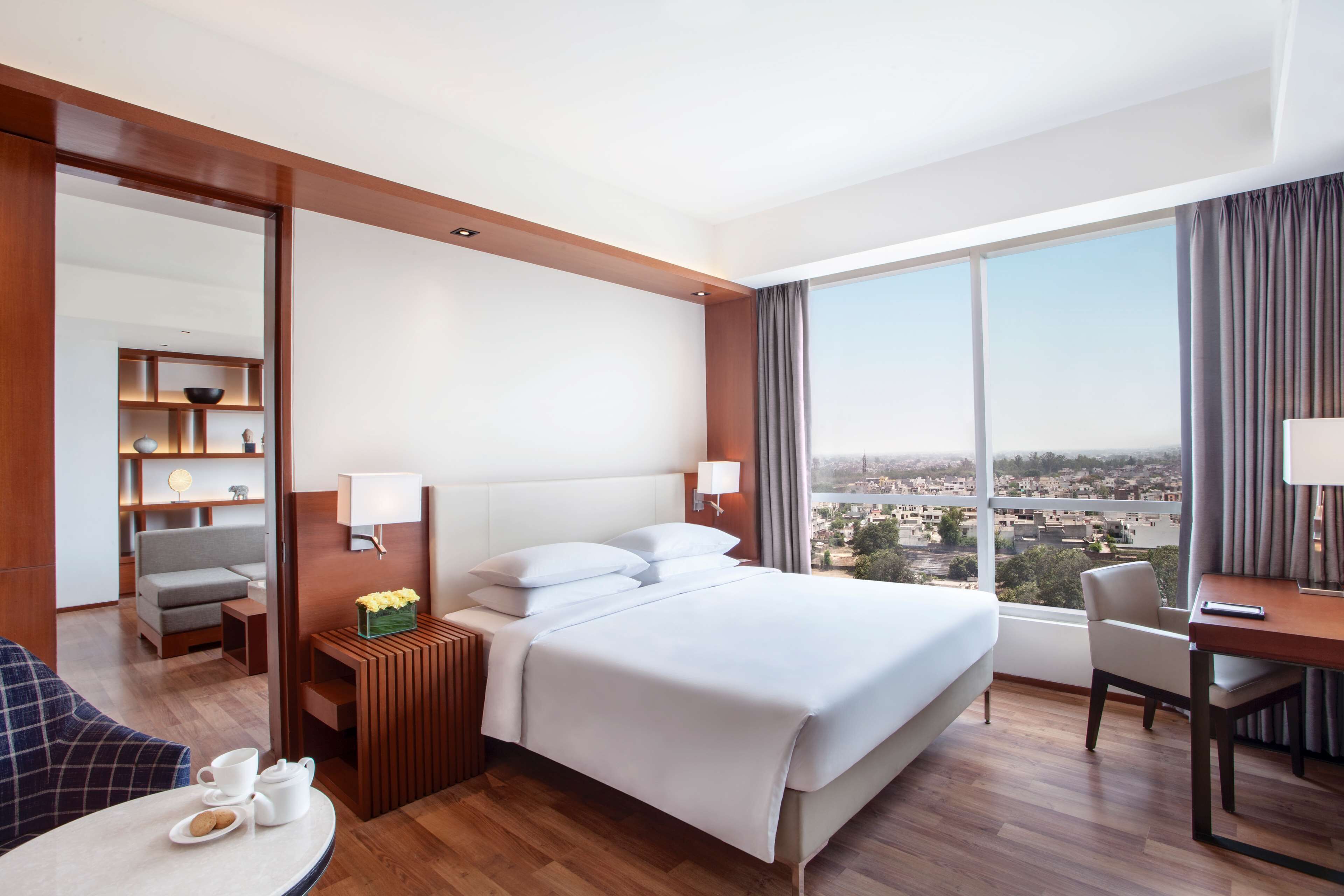 Hotelkamer met een groot bed, bureau, stoel, een klein tafeltje met kop en schotel, en uitzicht op de stad door grote ramen.