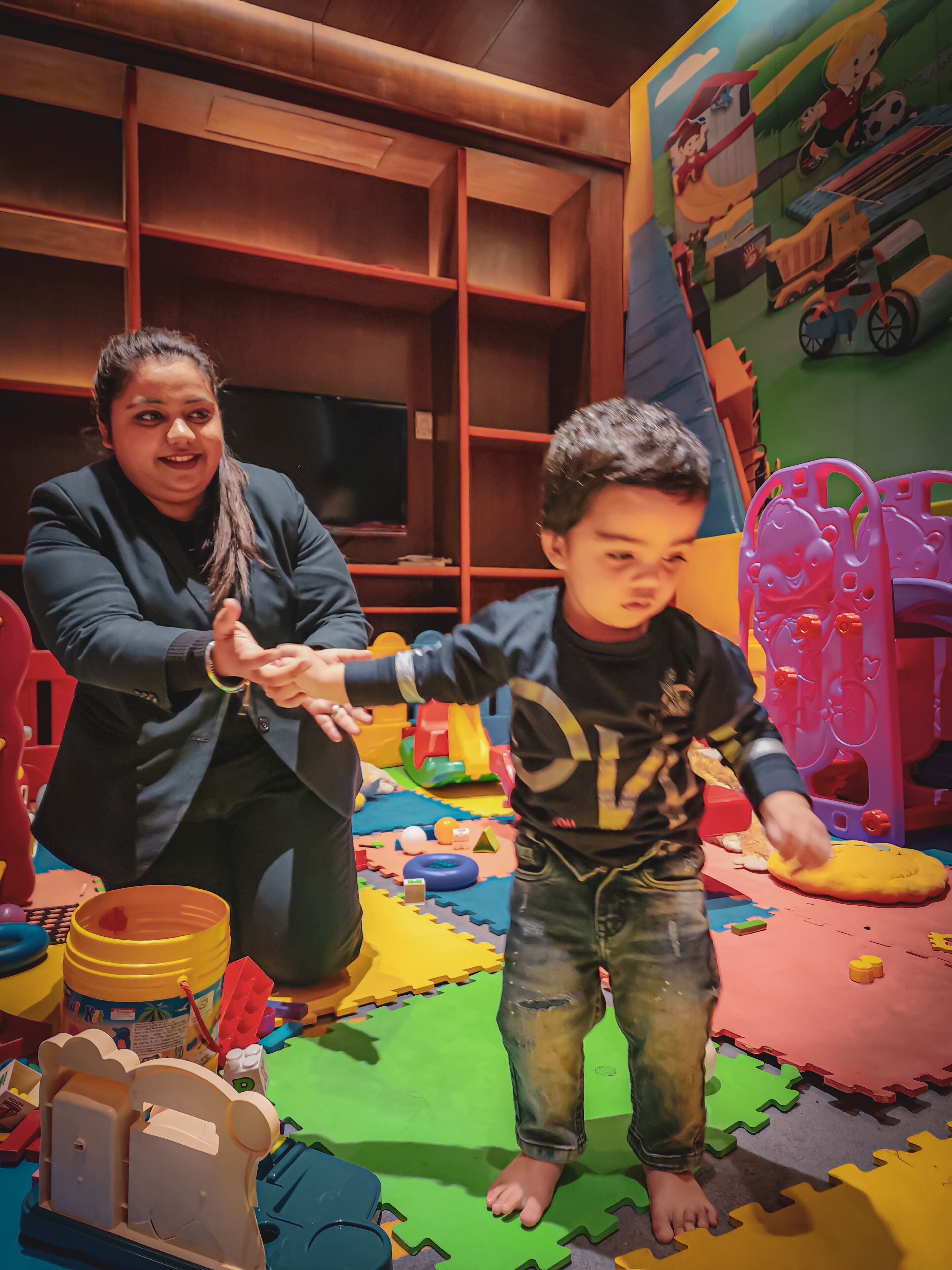 Un enfant et un adulte dans un espace de jeux équipé de tapis en mousse colorés, de jouets et d’une structure de jeu.