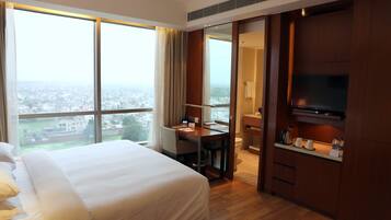 A hotel room with a large bed, a desk with a chair, a television, and a view of the city through large windows.