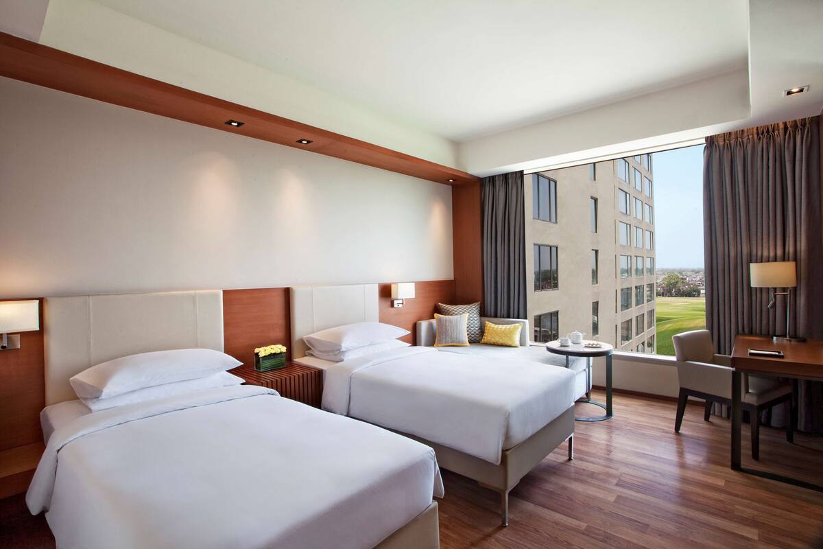 A hotel room with two beds, a desk, a chair, and a view of a building through large windows.