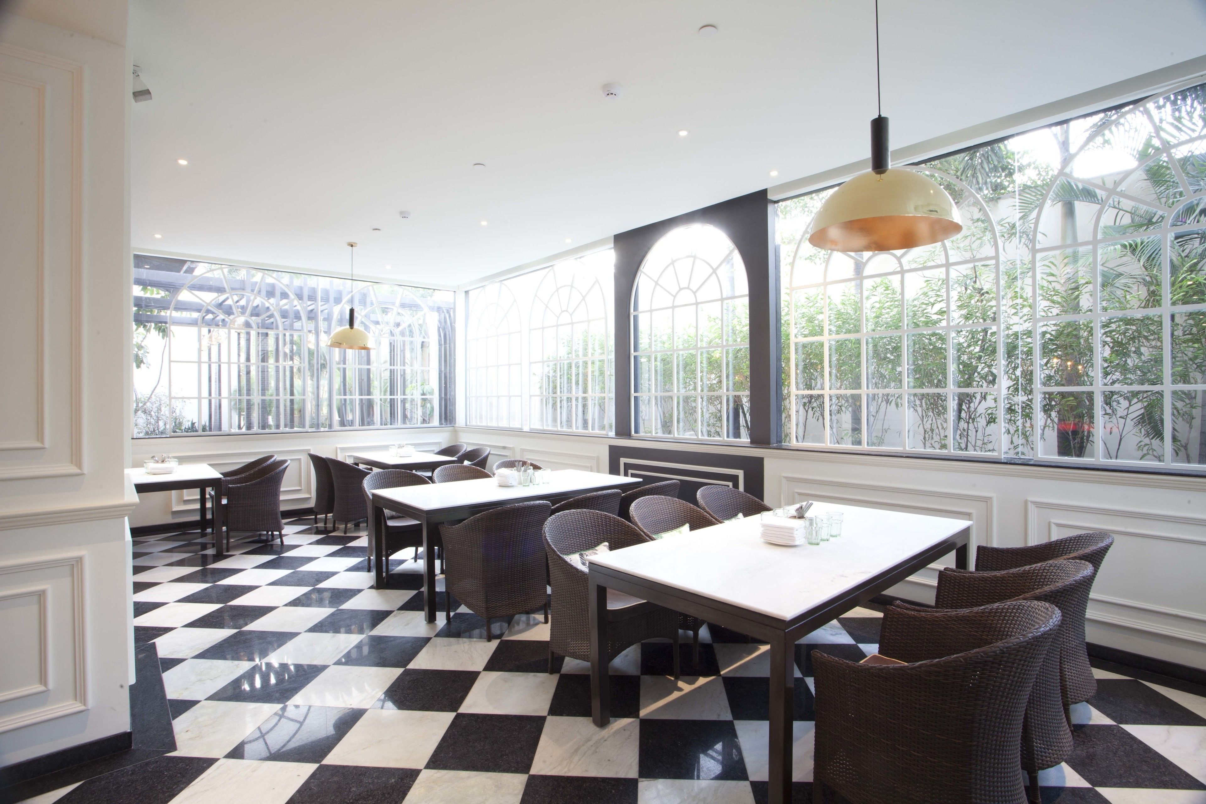 A restaurant with checkered flooring, white walls, and large windows offering a view of greenery. Tables and wicker chairs are arranged for dining.