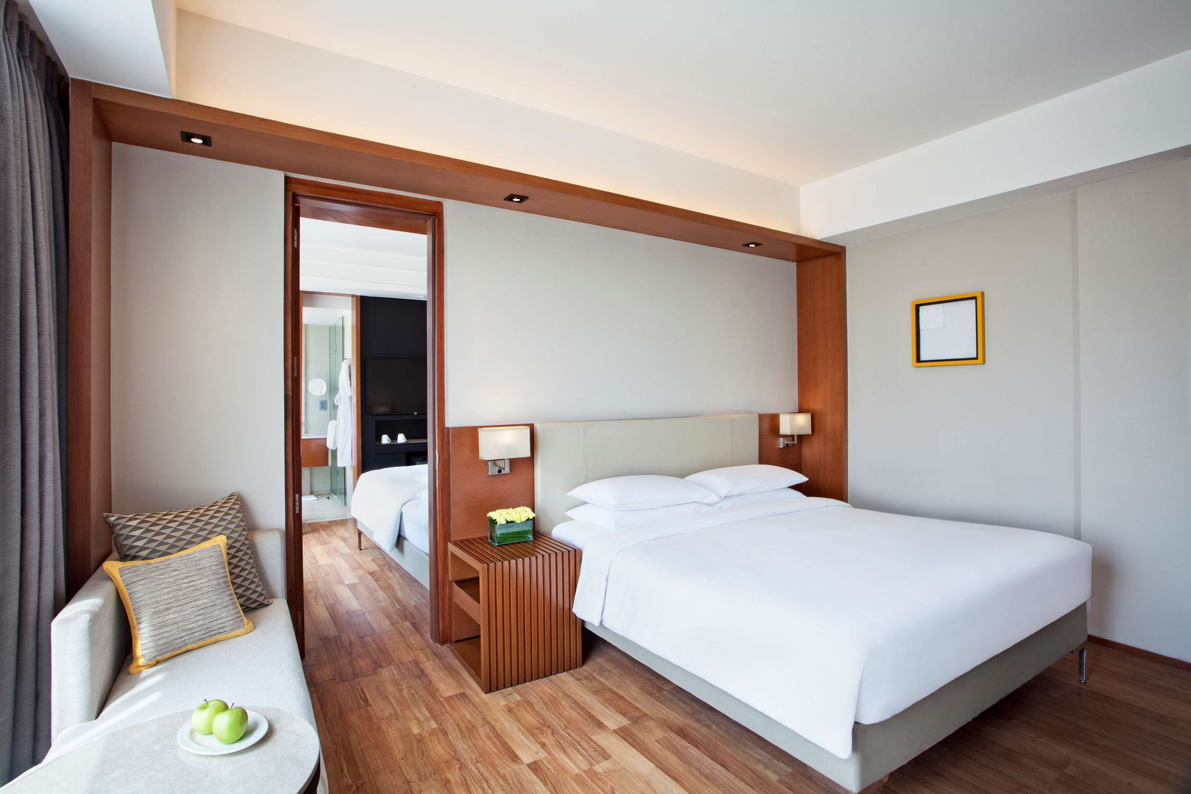 Habitación de hotel con cama, mesita de noche con lámpara, sofá con cojín y una pequeña mesa con manzanas.